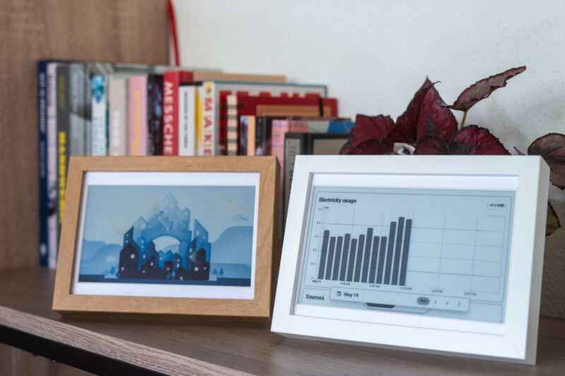 Two OpenDisplay e-paper art frames on a desk, with a row of books and a plant behind them. The color art frame on the left is in a wooden frame and shows a Home Assistant illustration made by Clelia Rella. The mono art frame on the right is showing a dashboard that shows current electricity usage.